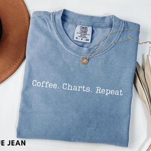 Puede incluir: Camiseta azul vaquero con el texto blanco "Coffee. Charts. Repeat". La camiseta tiene cuello redondo y mangas cortas. Un collar dorado con un colgante está drapeado sobre la camiseta. Un sombrero marrón y una planta seca están en el fondo.