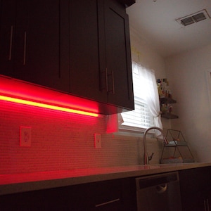 May include: A kitchen scene featuring dark brown cabinets with a red LED light strip illuminating the backsplash. The backsplash is a light-colored, textured tile. A window with a white curtain is visible, along with a stainless steel faucet and a dishwasher.