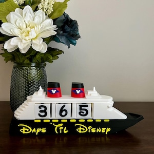 Könnte beinhalten: Ein schiffsförmiger Countdown-Kalender in Weiß und Schwarz mit dem Text "Days Til Disney" in Gelb. Der Kalender zeigt "365" auf rotierenden Blöcken. Zwei rot-schwarze Schornsteine ​​zeigen ein Mickey Mouse-Design.