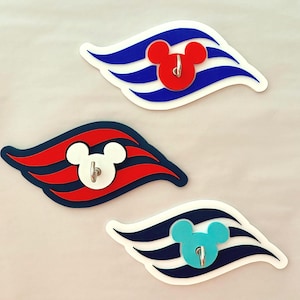 May include: Three decorative wall hooks featuring a Mickey Mouse head design. Each hook has a different color scheme: red and blue, red and white, and blue and white. The hooks are shaped like a stylized flag.