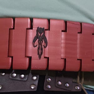 May include: A red, segmented belt with a black Mandalorian symbol on each segment. The belt is worn around a person's waist.