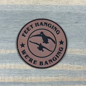 May include: A circular brown leather patch featuring a black engraved design of two flying ducks in the center. The text "FEET HANGING WE'RE BANGING" is printed in a circular black font around the ducks, with two black stars separating the phrases.