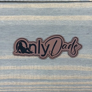 May include: A brown sticker with a black outline and black text that reads Only Dads. The letter O is replaced by a black silhouette of a person wearing a baseball cap and reclining on their side, integrated into the sticker design.