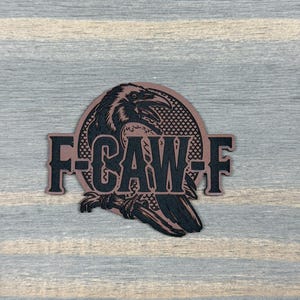 May include: A circular patch featuring a black illustration of a raven perched on a branch against a textured background. The text F-CAW-F is printed in bold black letters across the center of the raven. The patch has a copper-colored border.