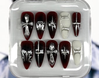 May include: A set of press-on nails in a clear plastic case. The nails feature a dark red base with silver cross and star designs. Some nails are solid white with silver accents. The text "areum nail studio" and "dominATE Stray Kids Inspired Nails" are visible.