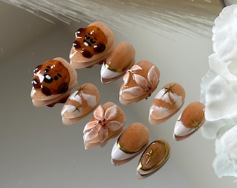 May include: Set of ten nude-colored press-on nails with various designs. Some nails feature a brown cartoon bear face, while others have white and gold floral designs.