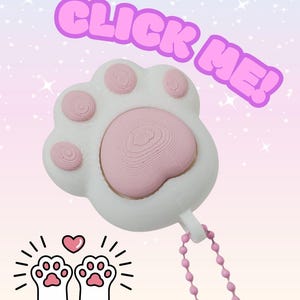 Peut inclure: Un charm en forme de patte de chat blanc et rose avec une chaîne perlée rose. Le charme porte le texte "CLICK ME!" en lettres roses à bulles. En dessous, deux pattes de chat avec un cœur.