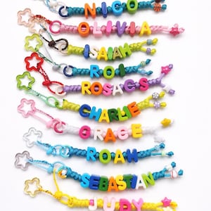 May include: A collection of colorful name keychains. Each keychain features a different name spelled out with vibrant letter beads. The keychains are made with braided cords and adorned with star-shaped charms and small decorative beads.
