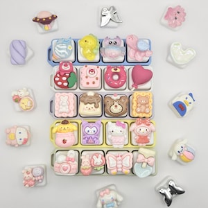 Peut inclure: Une collection de petites breloques décoratives en résine colorée représentant divers personnages de dessins animés, animaux, aliments et formes. Les breloques sont disposées dans cinq plateaux en plastique et éparpillées sur une surface blanche.