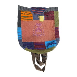 May include: A patchwork backpack featuring a central terracotta panel with an embroidered owl design. The bag is constructed from various colored fabric squares including orange, blue, green, and maroon, with distressed horizontal slits. A yellow tag is attached.