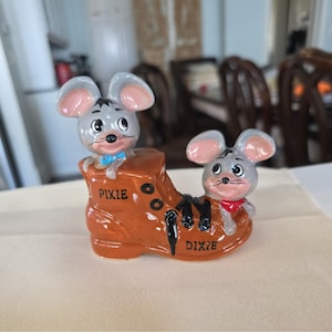 May include: A ceramic figurine featuring two cartoon mice in a brown boot. One mouse is wearing a blue bow tie and the other a red scarf. The boot has the words "PIXIE" and "DIXIE" on it. The mice are gray with pink ears.