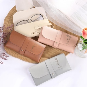 Pode incluir: Três estojos para óculos personalizados com designs florais e nomes. Os estojos são marrons, rosa e cinza.