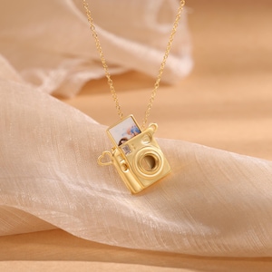 Könnte beinhalten: Goldene Halskette in Form einer Kamera mit einem kleinen Fotoeinsatz. Die Halskette hat einen herzförmigen Anhänger an der Seite.