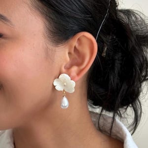 May include: White flower-shaped earrings with a pearl drop. The earrings are worn on the ear. The earrings have a gold-colored post. The earrings are a floral design. The earrings are a fashion accessory.