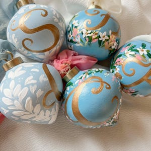 May include: Five light blue Christmas ornaments with gold-colored initials and floral designs. The ornaments have gold-colored caps and white ribbons. Some ornaments feature the letter "S" and others feature the letter "C".