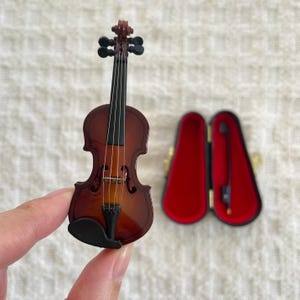 Puede incluir: Un viol&iacute;n en miniatura con un cuerpo de madera marr&oacute;n y clavijas negras. El viol&iacute;n est&aacute; en una caja de terciopelo rojo.