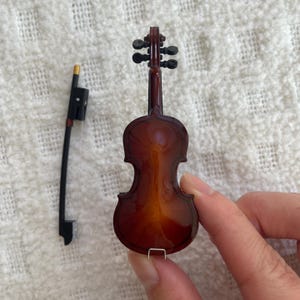 Puede incluir: Un viol&iacute;n en miniatura con un cuerpo de madera marr&oacute;n y clavijas negras. El viol&iacute;n est&aacute; sobre una superficie blanca y est&aacute; siendo sostenido por una mano.