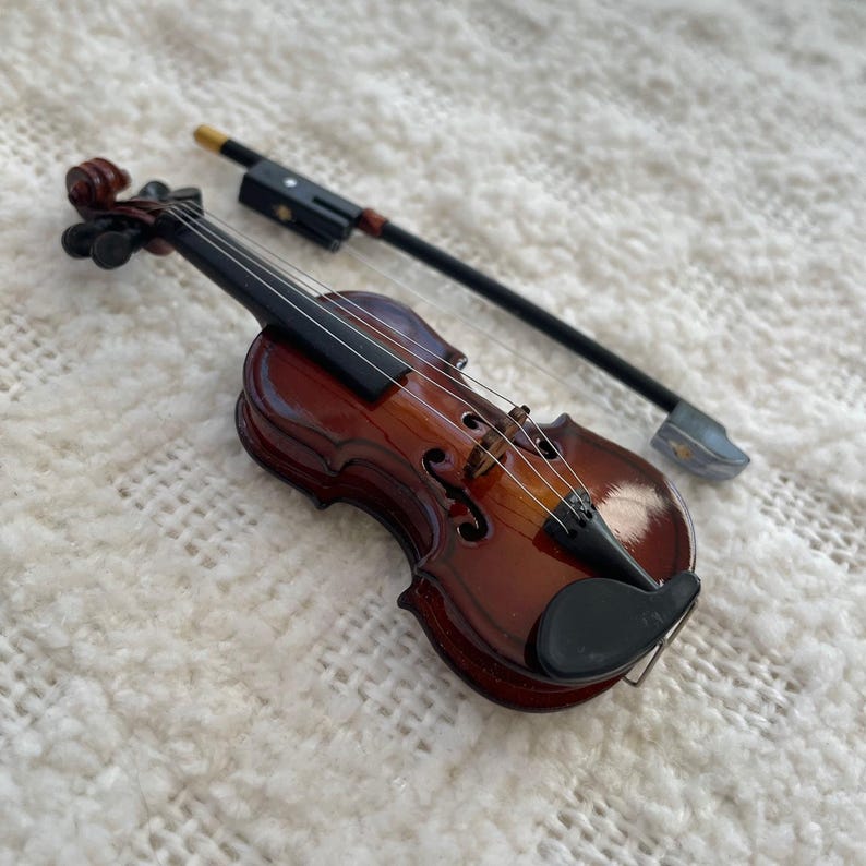 Puede incluir: Un viol&iacute;n en miniatura con un cuerpo de madera marr&oacute;n y cuerdas negras, y un arco negro a juego.