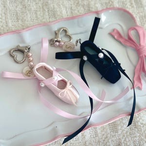 以下が含まれることがあります： パールがあしらわれたリボン付きのバレーシューズチャーム2個。ピンクのシューズチャームには「E」の文字が、黒いシューズチャームには「S」の文字が書かれています。