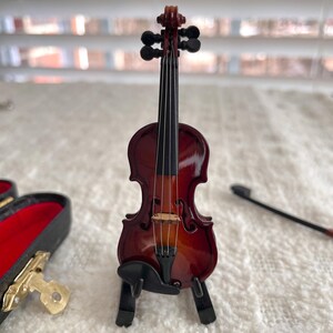 Puede incluir: Un viol&iacute;n en miniatura con un acabado rojo-marr&oacute;n oscuro, presentado en un soporte negro. El viol&iacute;n tiene un arco negro y es un art&iacute;culo decorativo.