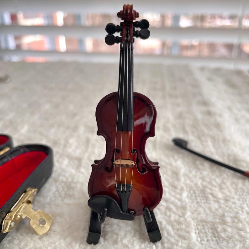 Puede incluir: Un viol&iacute;n en miniatura con un acabado rojo-marr&oacute;n oscuro, presentado en un soporte negro. El viol&iacute;n tiene un arco negro y es un art&iacute;culo decorativo.