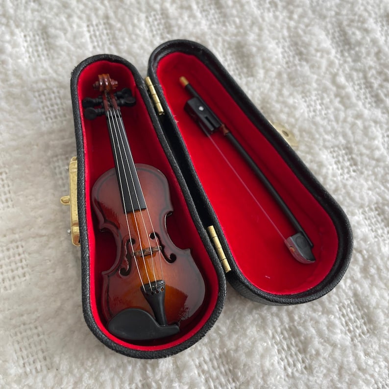 Puede incluir: Un viol&iacute;n en miniatura con un cuerpo de madera marr&oacute;n y cuerdas negras en una caja forrada de terciopelo negro con interior rojo. El viol&iacute;n viene con un arco negro.