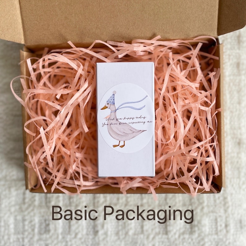 Puede incluir: Una peque&ntilde;a caja blanca con una pegatina de un pato de dibujos animados que lleva un sombrero azul y blanco. La pegatina dice "Te deseo un feliz d&iacute;a, me has estado esperando". La caja est&aacute; sentada en una cama de papel de seda rosa.