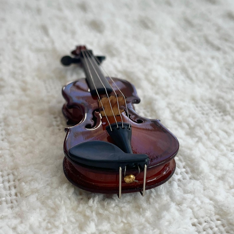 Puede incluir: Un peque&ntilde;o viol&iacute;n de madera con un acabado marr&oacute;n oscuro y un reposacabezas negro. El viol&iacute;n tiene cuatro cuerdas y est&aacute; sobre una superficie blanca y esponjosa.