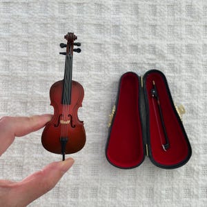 以下が含まれることがあります： 茶色の木製のボディと黒い弦を持つミニチュアチェロ。赤いベルベットの内装の黒いケースに入っています。