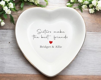Könnte beinhalten: Eine herzförmige weiße Keramikschale mit goldenem Rand. Auf der Schale steht der Text "Sisters make the best friends" mit einem roten Herz und den Namen "Bridget & Allie". Sie liegt auf einer Holzoberfläche.