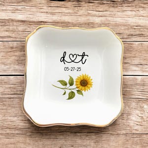 Peut inclure: Petit plat à bijoux en céramique blanche avec un bord doré et un motif de tournesol. Le plat porte les initiales "d♡t" et la date "05-27-25" en écriture noire. Le tournesol est jaune et marron avec des feuilles vertes.