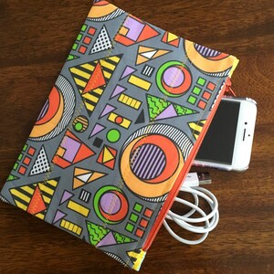 May include: A gray zippered pouch with a geometric pattern in orange, yellow, green, purple, and white. The pouch is open and a white charging cable and a black smartphone are inside.