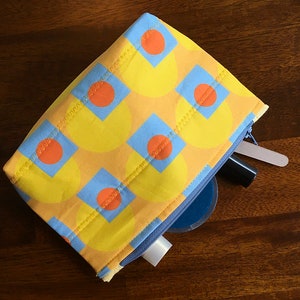 May include: A yellow and blue geometric patterned zippered pouch with a blue zipper. The pouch is open and contains a white tube, a blue container, and a black tube.