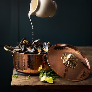 Puede incluir: Una olla de cobre martillada con una tapa con un adorno de cabeza de león. La olla está llena de mejillones y se está vertiendo una salsa blanca sobre ellos.