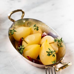 May include: A dish of poached pears, red chillies, and thyme sprigs. The pale yellow pears are in a copper-coloured oval dish with gold handles. A fork and knife are in the foreground. The dish is on a marble surface.