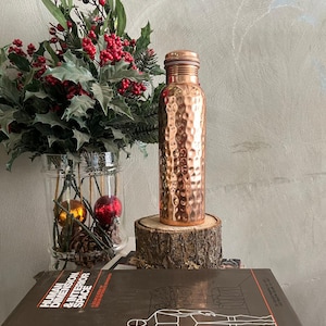 May include: A cylindrical copper water bottle with a hammered, dimpled texture and a matching screw-top lid. The bottle stands upright on a circular wooden log slice, positioned in front of a glass vase filled with green leaves and red berries.