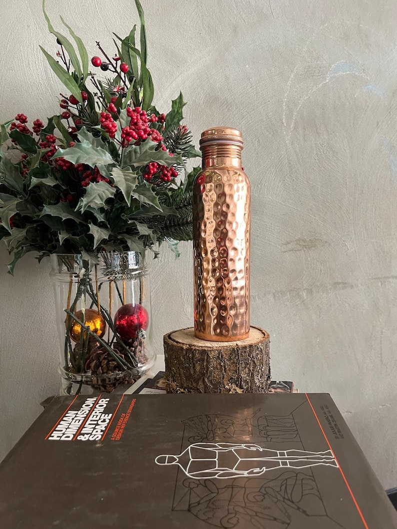 May include: A cylindrical copper water bottle with a hammered, dimpled texture and a matching screw-top lid. The bottle stands upright on a circular wooden log slice, positioned in front of a glass vase filled with green leaves and red berries.
