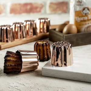 May include: Copper-colored canelé molds and baked canelés are displayed on a white surface. The molds have a fluted design, and the baked canelés have a dark, caramelized exterior. Ingredients like flour and eggs are visible in the background, suggesting a baking theme.