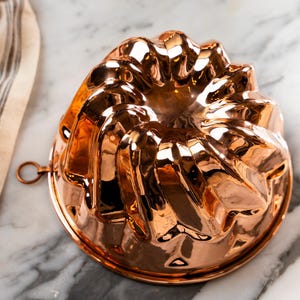 May include: A shiny copper bundt cake pan with a fluted design. The pan has a small loop handle on the side.