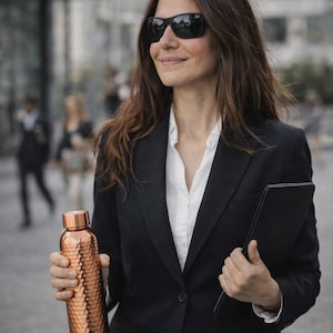 May include: A metallic copper water bottle with a textured, hammered surface and a smooth, cylindrical cap. The bottle features a reflective finish and a tapered neck, designed for holding liquids, held in a hand alongside a black folder.