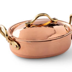 May include: An oval copper casserole dish with brass handles and a lid. The dish is shiny and has a classic design.