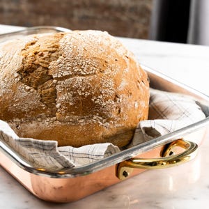 Puede incluir: Una hogaza de pan redondo y crujiente descansa en una fuente de horno color cobre con asas doradas. El pan está sobre un paño a cuadros, en una fuente sobre una superficie blanca.