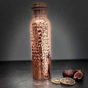 May include: A cylindrical copper water bottle featuring a textured, hammered exterior finish and a matching metallic screw-top lid. The bottle has a narrow neck and a smooth, flat base, reflecting light across its polished, warm-toned metallic surface.