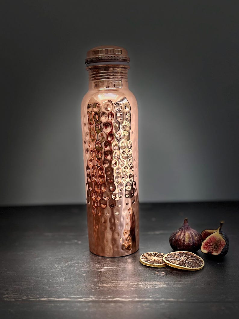 May include: A cylindrical copper water bottle featuring a textured, hammered exterior finish and a matching metallic screw-top lid. The bottle has a narrow neck and a smooth, flat base, reflecting light across its polished, warm-toned metallic surface.