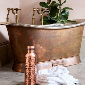 May include: A hammered copper water bottle with a screw-top lid stands next to a stack of folded white towels. Behind them is a large copper bathtub with brass faucets and a metal plaque reading Catchpole & Rye Kent England.