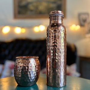May include: A hammered copper water bottle with a matching hammered copper tumbler. Both items feature a reflective, textured surface with a metallic finish. The bottle has a cylindrical shape with a screw-top lid, while the tumbler is rounded.