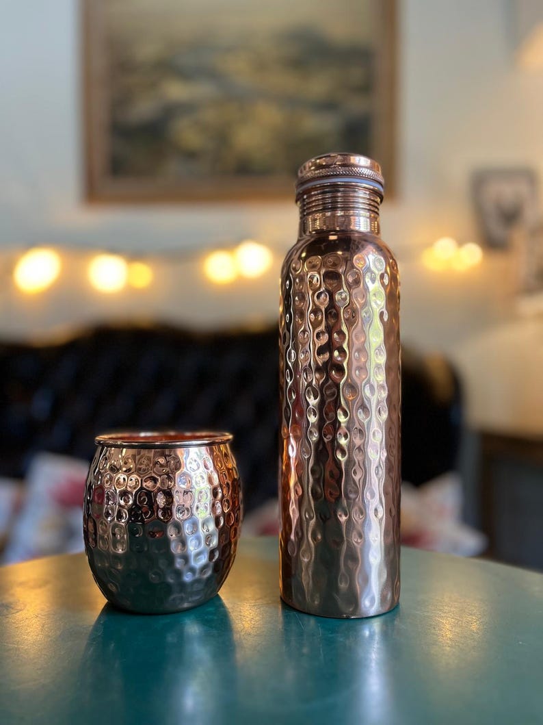 May include: A hammered copper water bottle with a matching hammered copper tumbler. Both items feature a reflective, textured surface with a metallic finish. The bottle has a cylindrical shape with a screw-top lid, while the tumbler is rounded.