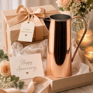 May include: A polished copper pitcher with a tall cylindrical body and a curved handle, presented in a gift box with a beige ribbon. A tag attached to the ribbon reads Copper Anniversary with a heart, and a card reads Happy Anniversary with a heart.