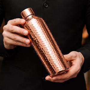 May include: A cylindrical copper water bottle featuring a distinct hammered texture across its entire surface. The bottle includes a matching smooth copper screw-top cap. The metallic finish reflects light, highlighting the indented pattern.