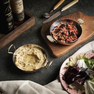 May include: A food still life featuring two small metal pans with gold handles, one filled with hummus and the other with a tomato and aubergine dish. A wooden board, salad, and bottles of oil are also present.